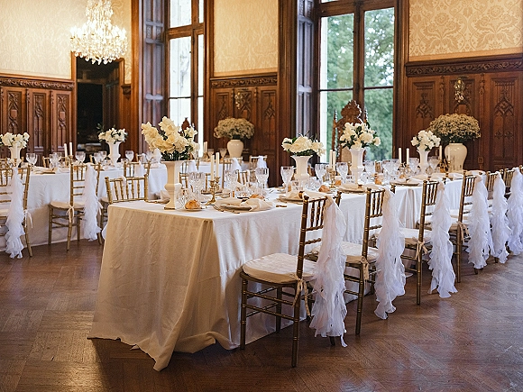 Reception tablescape with white wedding tablescape details, tall white floral centerpieces, taper candles, and gold Chiavari chairs under a chandelier