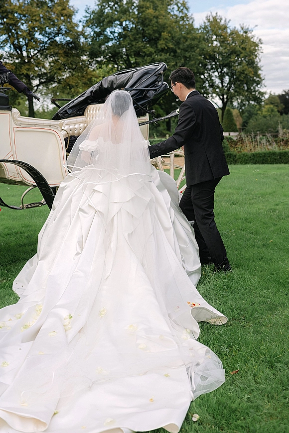 Wedding exit as groom in tuxedo helps bride with cathedral veil and train into a horse-drawn carriage on a green lawn