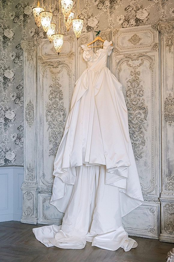Wedding dress hanging from a dress hanger, off the shoulder ball gown with long train against ornate paneled wall and pendant lights