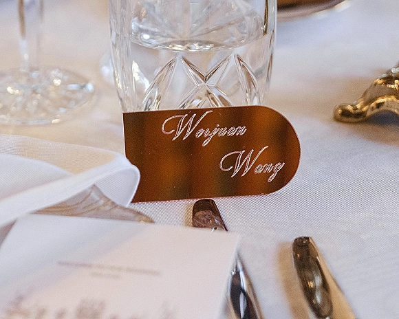 Wedding place card in copper with calligraphy lettering set on a cut crystal water glass beside a white napkin and silver flatware on white linen