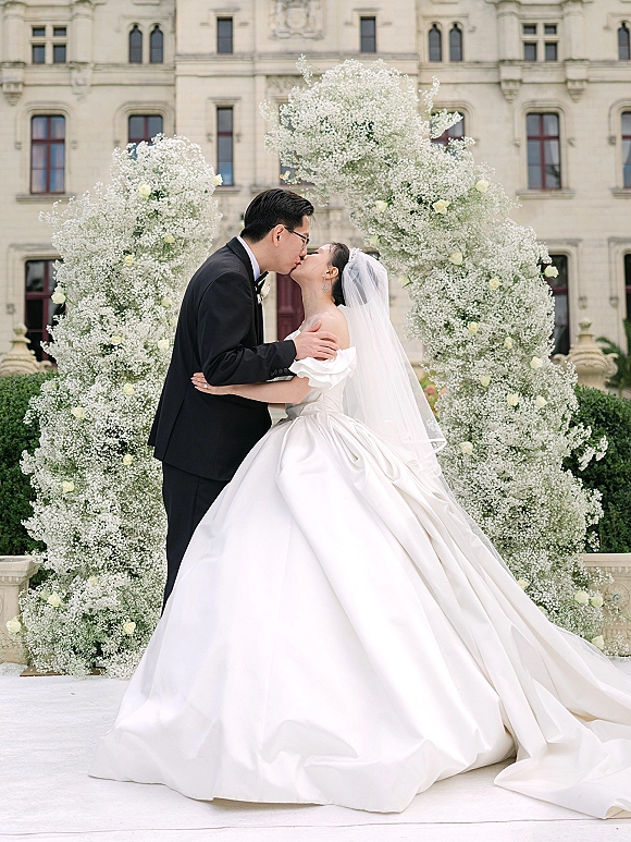 Wedding kiss portrait of bride and groom kiss under a white floral arch, her ball gown and veil flowing before a stone mansion facade