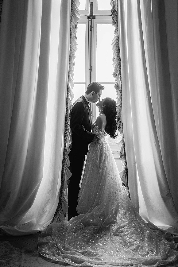 Wedding couple portrait in a black and white wedding portrait, bride and groom kissing by a tall window, backlit through curtains on wood floor