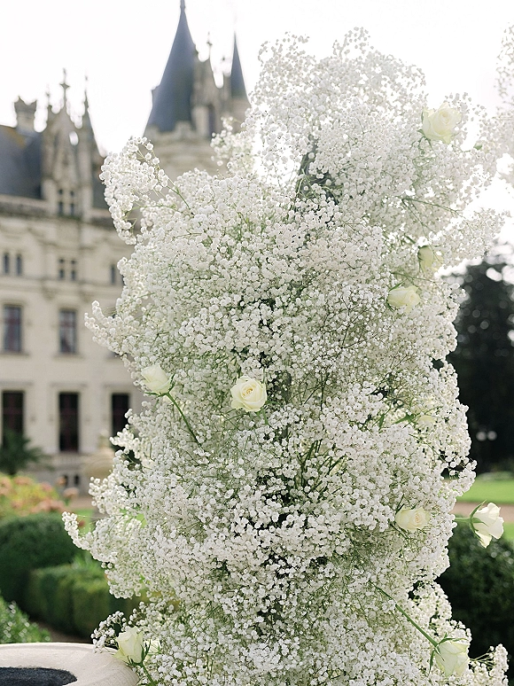 Wedding floral arrangement with baby’s breath and white roses on a garden lawn, set against a castle exterior and clipped hedges under sky