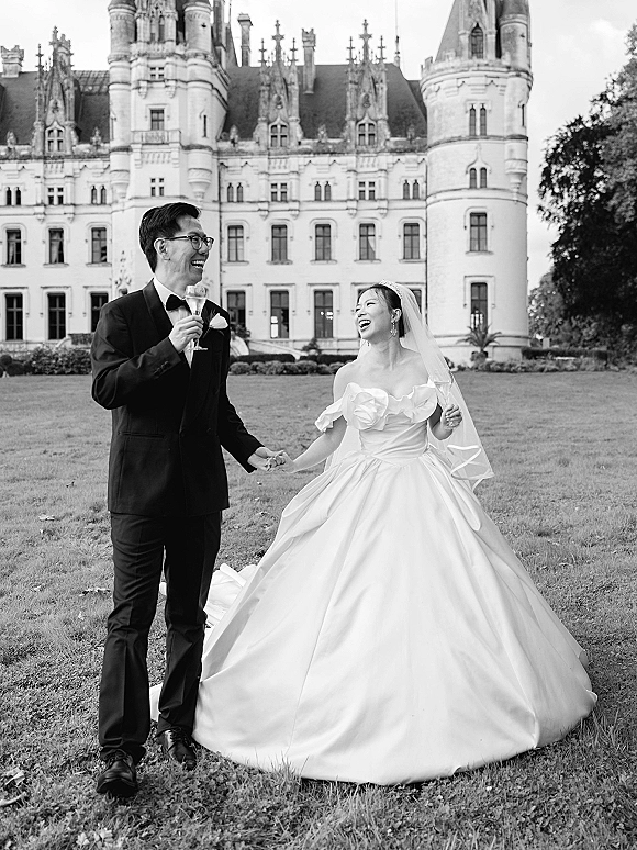 Couple portrait of bride and groom laughing, holding hands with a champagne glass, outside a castle on the lawn in black and white