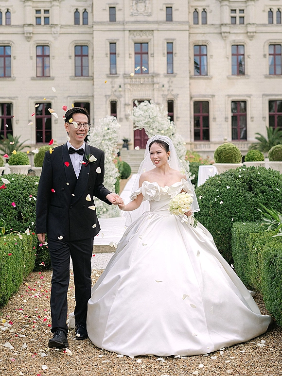 Recessional moment as bride and groom walk hand in hand through a rose petal toss, bouquet and veil flowing along a garden gravel path
