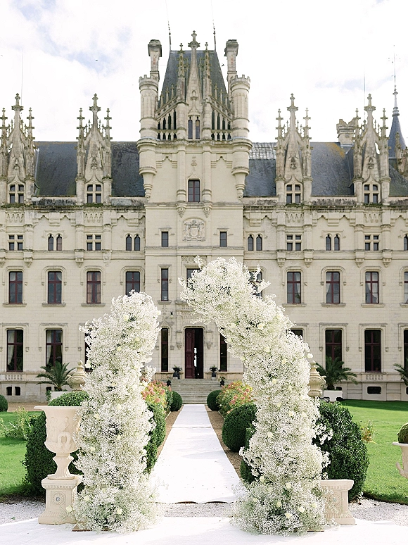 Wedding ceremony arch with a white floral arch of baby’s breath and roses framing a white aisle runner before a castle facade lawn