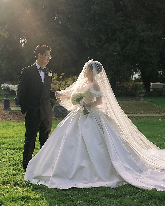 Couple portrait of bride and groom outdoors, her off-shoulder ball gown and bouquet, his tuxedo and glasses, on a sunlit garden lawn