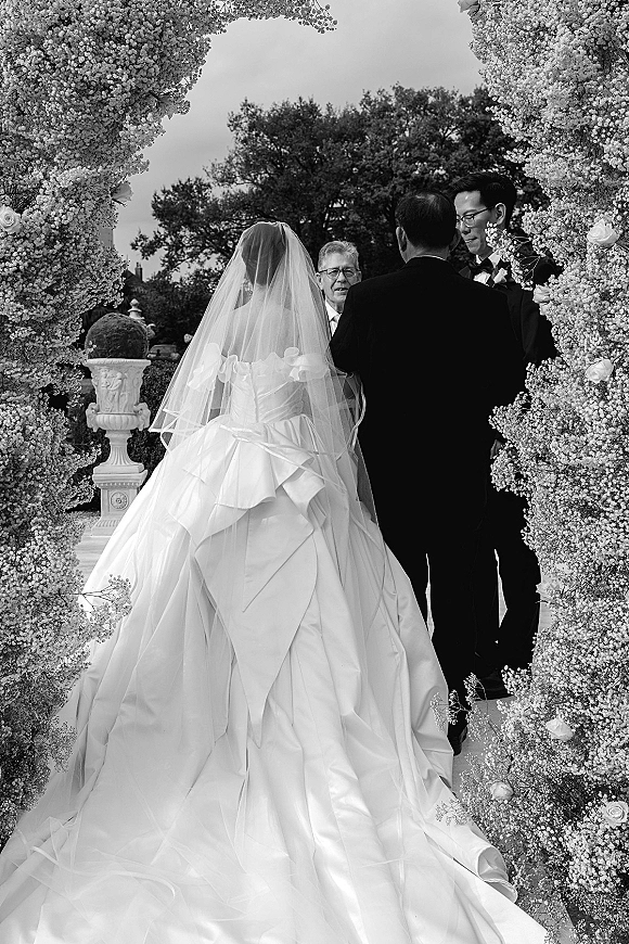 Wedding ceremony moment as bride in cathedral veil walks down aisle runner toward groom at floral arch in garden setting