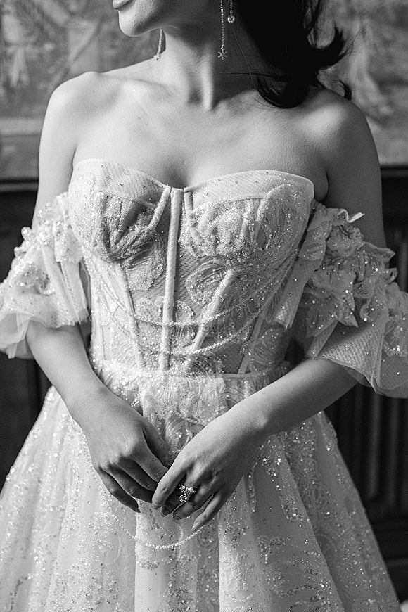 Wedding dress detail with off the shoulder neckline, glitter tulle and floral appliqués, bride’s hands showing engagement ring in window light