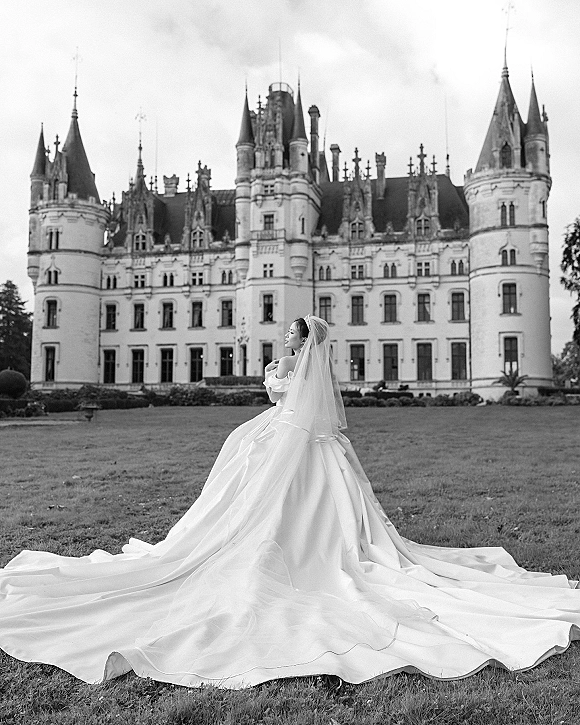 Bridal portrait of a bride in an off the shoulder gown with long train and veil, wearing a tiara on a castle lawn under cloudy skies