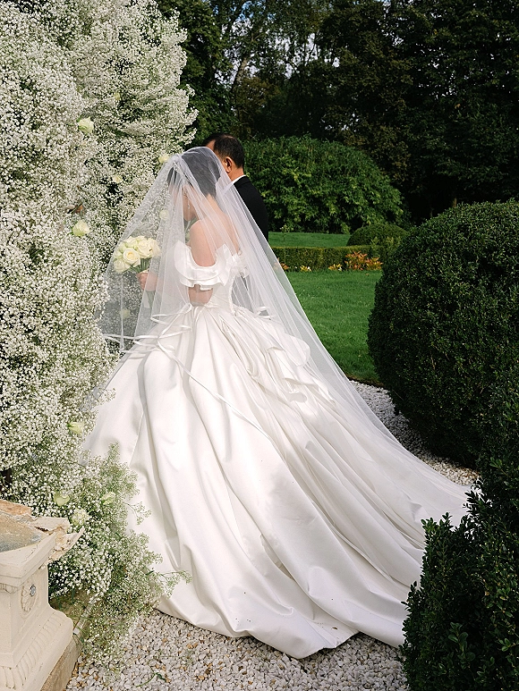 Couple portrait of bride and groom side profile in a garden, bride holding white rose bouquet with a flowing veil, groom in tuxedo