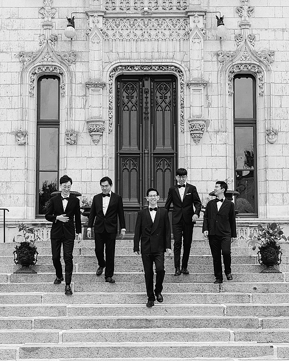 Groomsmen portrait of men in black tuxedos and bow ties walking down stone steps before ornate doors with arched windows
