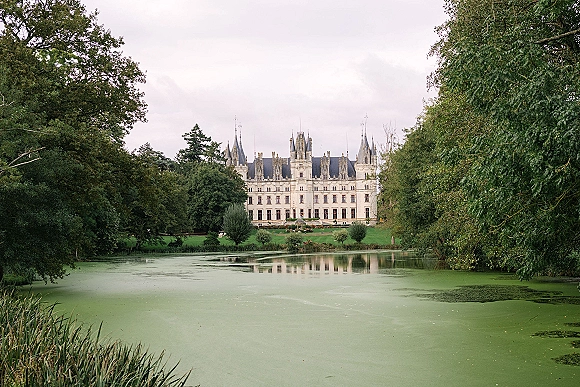 Castle wedding venue beside a calm pond, framed by trees and lawn under a cloudy sky, showcasing the chateau wedding venue exterior