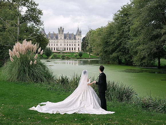 Couple portrait from behind, bride and groom holding hands on a green lawn by a lake with a castle under cloudy sky, wedding veil visible