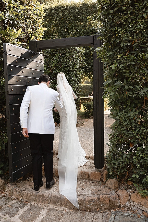 Couple portrait of bride and groom walking away, holding hands as her cathedral veil trails on sunlit stone steps by a black garden gate