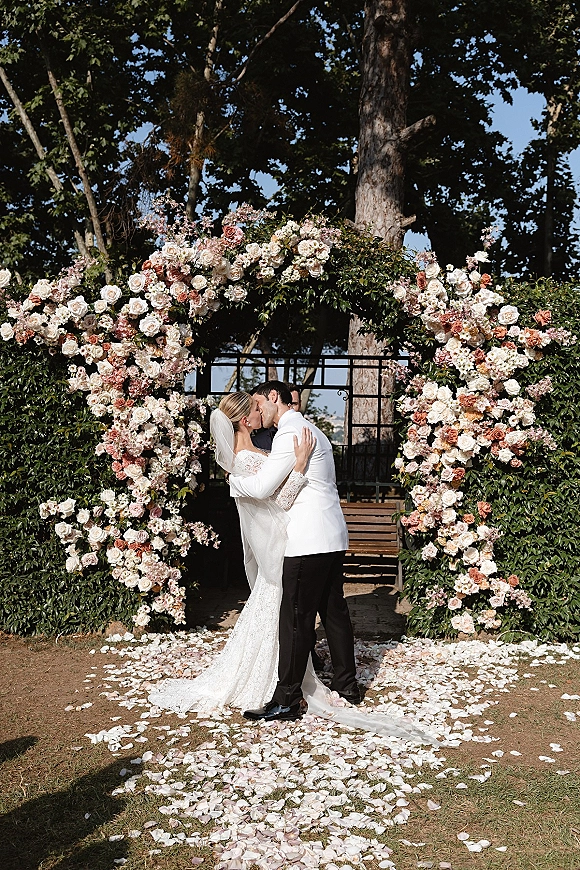 Wedding kiss portrait of bride and groom kissing beneath a rose and greenery floral arch, veil and lace gown beside a rose petal aisle