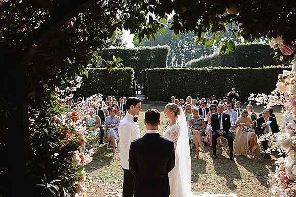 Ceremony moment as bride in lace dress and veil faces groom under a pink and white rose arch on a sunlit garden lawn