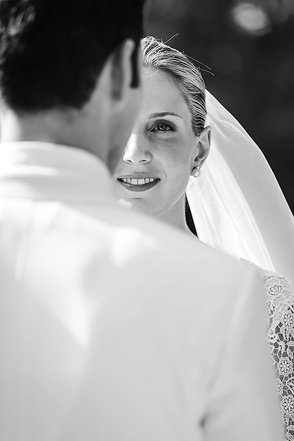 Bride portrait in black and white wedding portrait style, smiling at groom’s shoulder in foreground, veil and pearl earrings in soft greenery bokeh