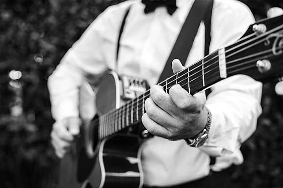 Wedding musician playing an acoustic guitar wedding set, wearing bow tie and suspenders with wristwatch against blurred outdoor greenery