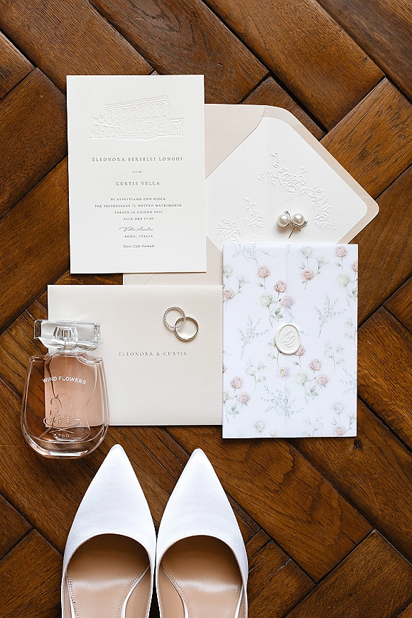 Invitation suite with embossed wedding invitation, wax seal, rings, pearl earrings, perfume and bridal shoes on a herringbone floor