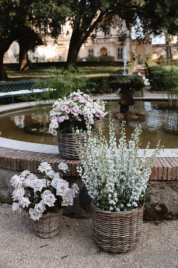 Wedding floral decor with wicker basket wedding flowers in pastel pink and white around a stone fountain in a garden courtyard setting