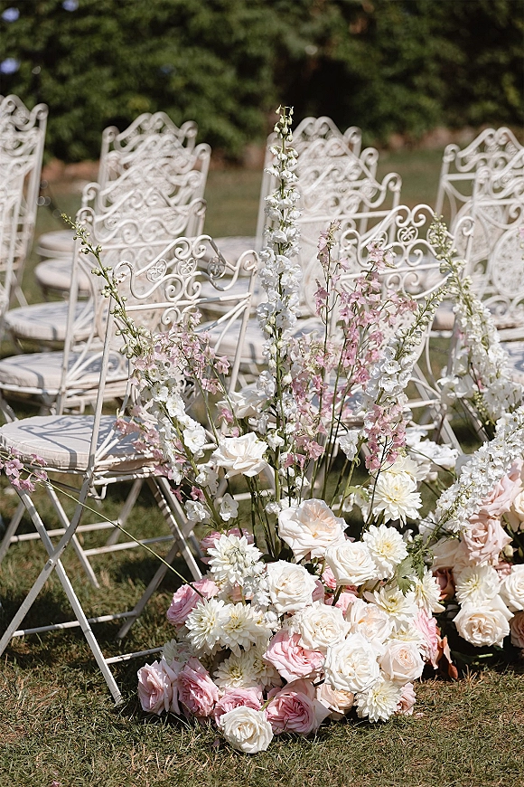 Ceremony aisle flowers with roses and dahlias in pastel pink, peach, and white with greenery beside white wrought iron chairs on a lawn