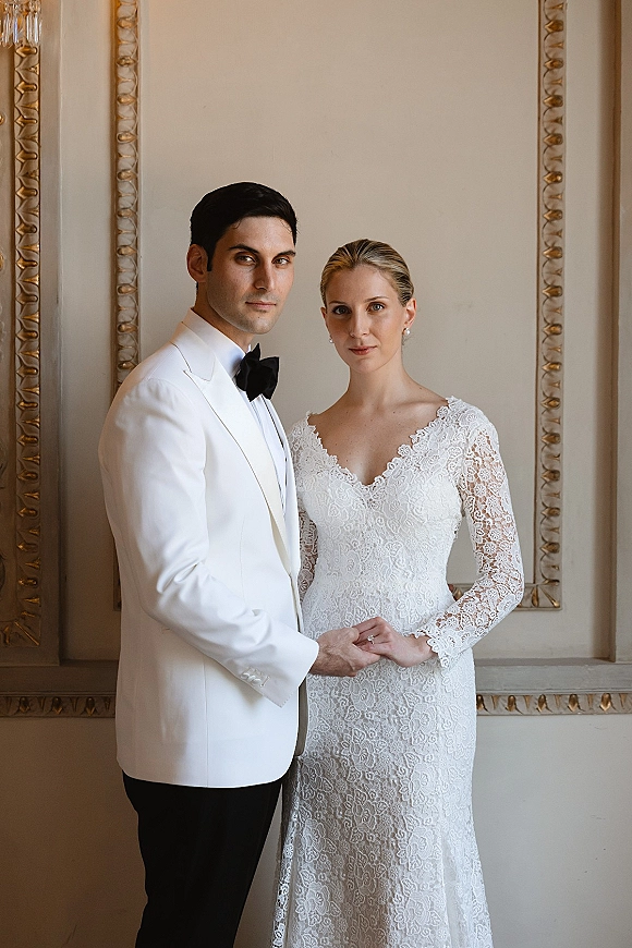 Couple portrait of bride and groom holding hands, showing lace long-sleeve gown and white tuxedo jacket against ornate wall molding
