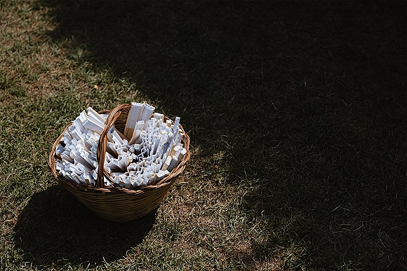 Wedding confetti toss setup with confetti toss packets in a wicker basket, filled with paper confetti, on a sunlit grass lawn with shadows