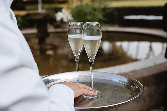 Champagne toast with wedding champagne glasses on a serving tray held by a server, set beside a pond and stone fountain edge outdoors