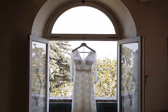 Wedding dress, lace wedding dress with long sleeves and v-neckline on a hanger by open arched window doors overlooking balcony and trees