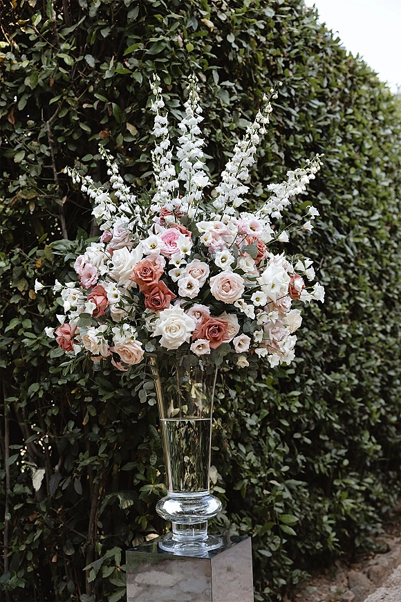 Wedding floral arrangement of roses, white blooms, and greenery in a tall clear glass vase on a pedestal against a hedge wall backdrop
