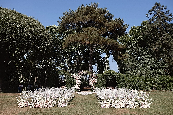 Ceremony setup for an outdoor wedding ceremony with a floral arch, white metal chairs, and flower petals lining the garden lawn aisle