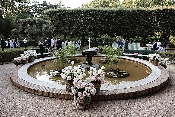 Garden cocktail hour scene with wedding cocktail hour decor around a round fountain with water lilies, potted roses, and guests mingling nearby