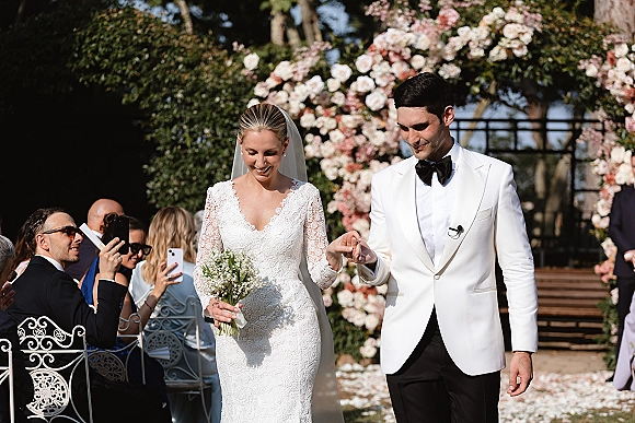 Wedding recessional as bride and groom walk the aisle holding hands, she in lace dress and veil with baby's breath bouquet under a floral arch