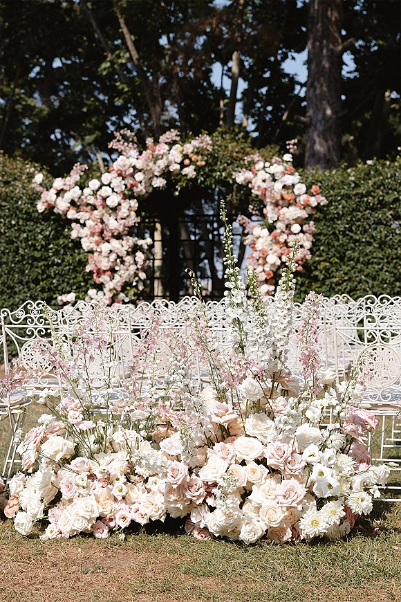 Ceremony backdrop with floral wedding arch of roses and tall stems, ground florals and white wrought iron chairs on a hedge-lined lawn