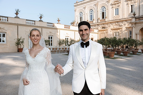 Couple portrait of bride and groom holding hands, bride in lace long-sleeve dress with veil, smiling in historic courtyard backdrop