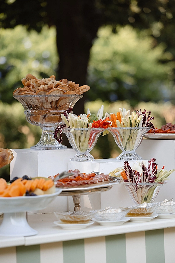 Wedding appetizer display with cocktail hour food table of charcuterie, crudité, fruit, and dips on glass pedestal bowls under trees