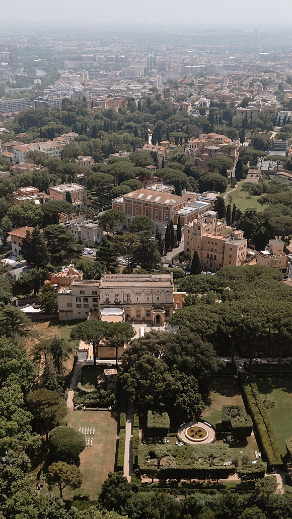 Wedding venue aerial view of a historic villa with manicured gardens, hedges and a fountain, bordered by trees with a city skyline beyond