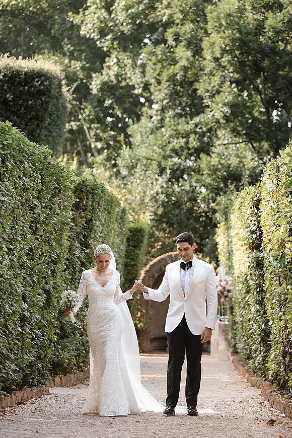 Wedding couple portrait of bride and groom walking hand in hand on a garden pathway, her lace long-sleeve gown and veil flowing