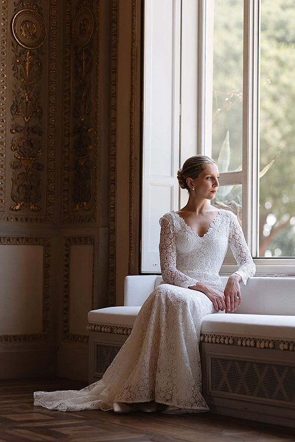 Bridal portrait of a bride in a lace long sleeve wedding dress, seated by a large window, looking out in natural light with greenery beyond