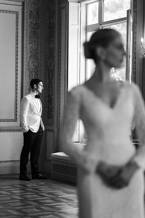 Wedding first look as groom waiting by window in white tux jacket and black bow tie, hands in pockets, bride enters behind in long-sleeve gown.