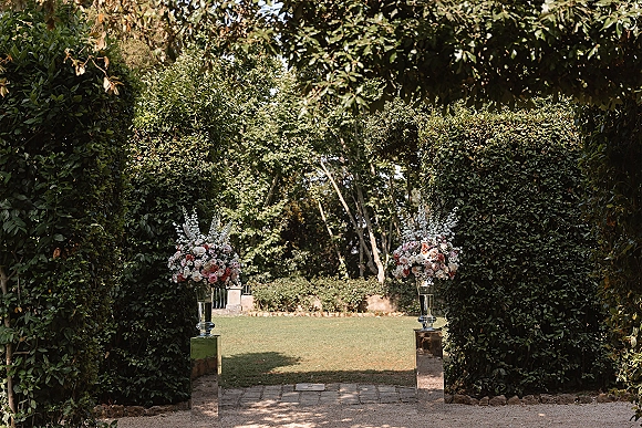Ceremony aisle entrance lined with rose bouquets in glass vases on pedestal stands, set along a gravel path amid garden hedges and trees