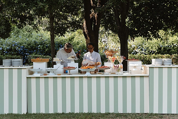 Wedding buffet table with sliced ham, bread rolls, and fruit garnish on tiered stands, set on a mint green striped counter in a garden lawn setting