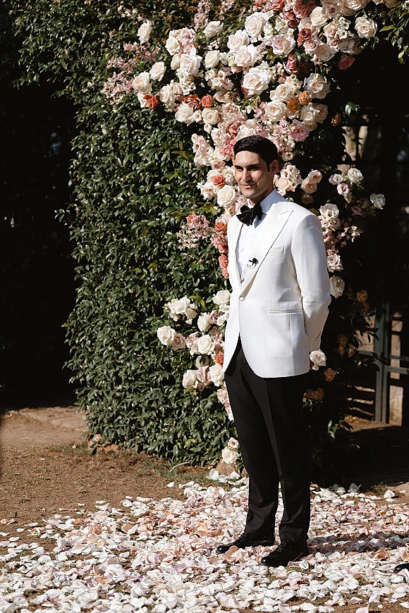 Groom portrait in a white tuxedo jacket with black bow tie, hands behind back by a floral arch with roses on an outdoor walkway