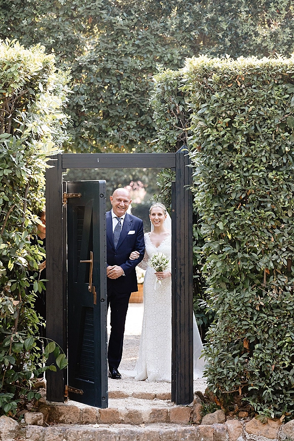 Processional moment as bride walking down aisle with her father, lace gown and veil, holding a white bouquet by an open garden gate
