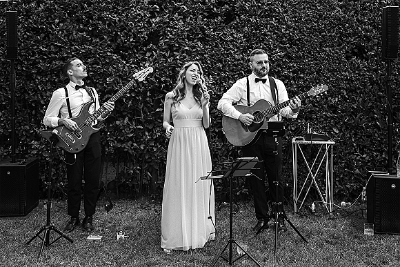 Wedding band performing live wedding music on a grass lawn, singer with microphone beside acoustic and bass guitars against a hedge wall