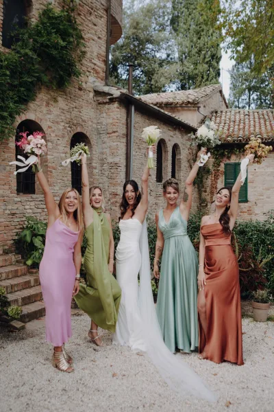 Bride with bridesmaids cheering in colorful bridesmaid dresses, raising bouquets with white ribbon wraps in an ivy brick courtyard
