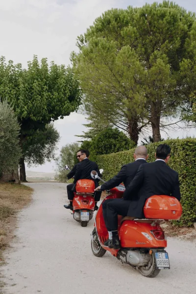 Wedding getaway with groomsmen on scooters riding red scooters in black suits and sunglasses along a country road under cloudy skies