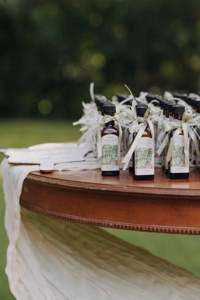 Wedding favors of mini liquor bottle favors with ribbon bows and custom labels arranged on a draped wooden table by greenery outdoors