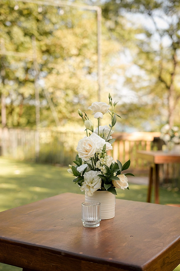 Wedding centerpiece with white rose centerpiece in a ceramic vase beside glass votive candles on a wooden table under market lights outdoors